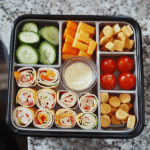 Overhead bento box with pinwheels, veggies, cheese cubes, crackers, and dip