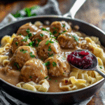 Close-up of Swedish meatballs in creamy gravy over egg noodles with a jam side and parsley